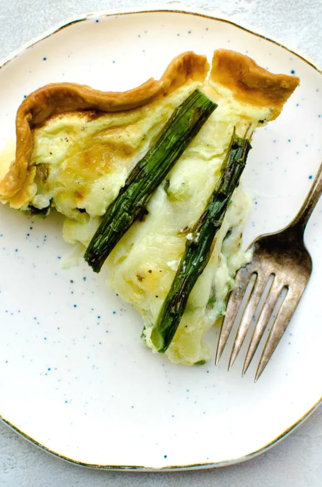 Asparagus quiche is a vegetarian quiche recipe perfect for a spring holiday brunch or weeknight dinner. Savory egg custard pie with asparagus and cheese. #asparagus #quiche #brunchrecipes #Easter slice of homemade asparagus quiche on small white plate