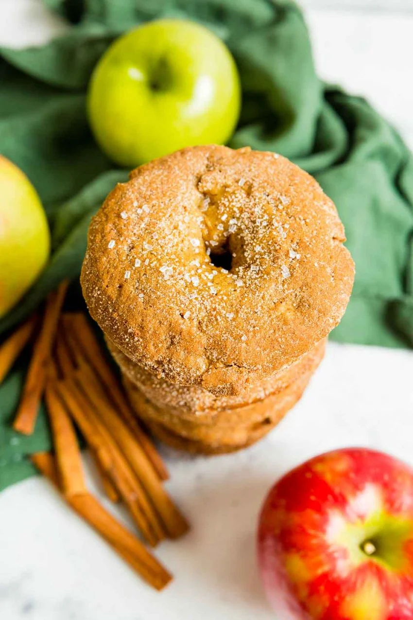 An overhead image of a stack of Apple cinnamon donuts with green and red apples, green napkin and cinnamon sticks in the background.