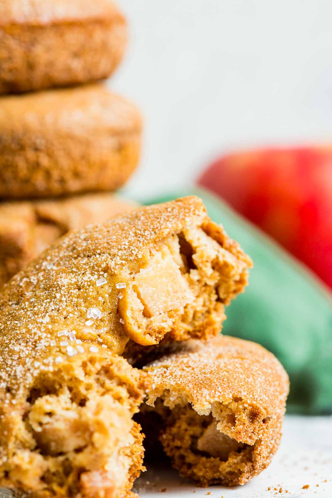 A close up of apple cinnamon donuts broken in half to show apple chunks. A stack of donuts with cinnamon-sugar, a red apple, green apple, cinnamon sticks and green cloth in the background.