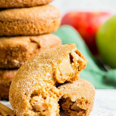 A close up of apple cinnamon donuts broken in half to show apple chunks. A stack of donuts with cinnamon-sugar, a red apple and green cloth in the background.