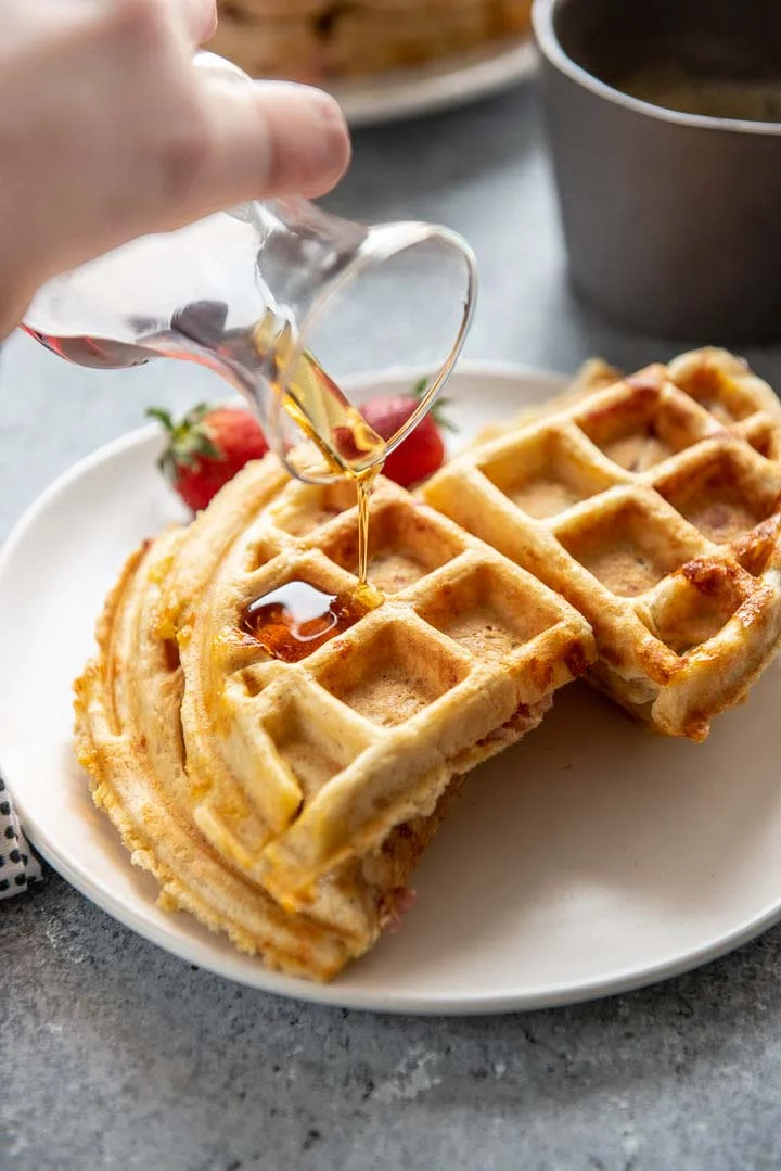 These savory waffles are a big hit for weekend brunch or weeknight dinner. They freeze well, so be sure to make a double batch! This stuffed waffles recipe uses leftover Easter ham and cheddar cheese. #waffles #leftoverham #brunch #recipes pouring syrup on savory waffles with ham and cheese