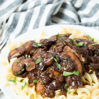 A tabletop view of chicken bourguignon topped with fresh parsley on a white plate and striped napkin in the background