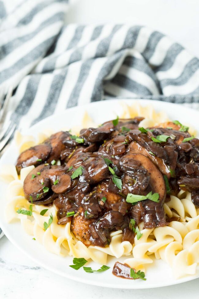 A tabletop view of chicken bourguignon topped with fresh parsley on a white plate and striped napkin in the background