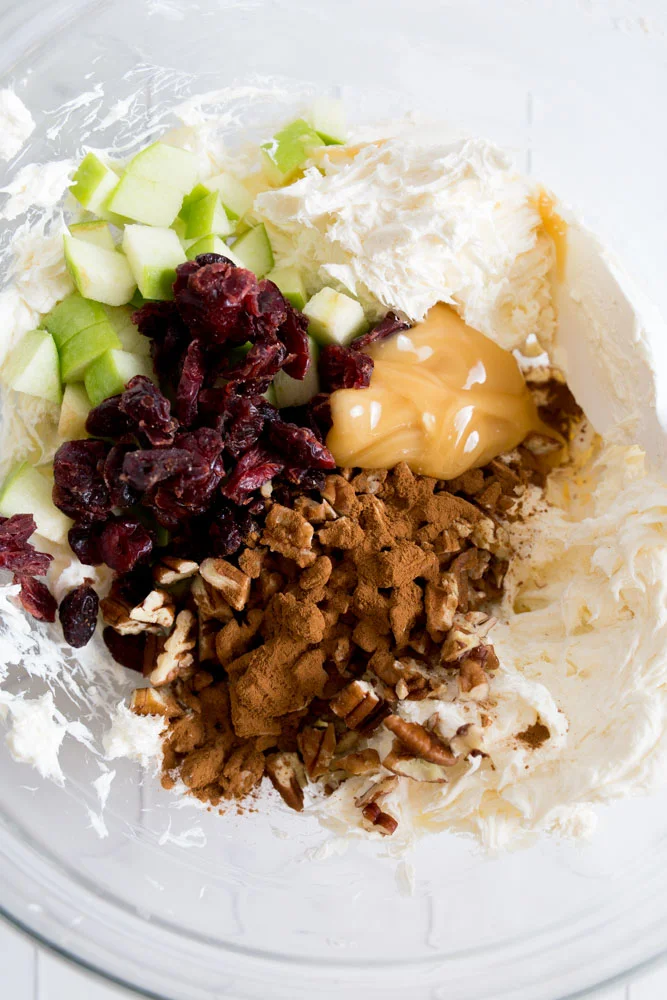 A close up image of a glass mixing bowl with cream cheese, dried cranberries, honey, nuts, cinnamon, and apples.