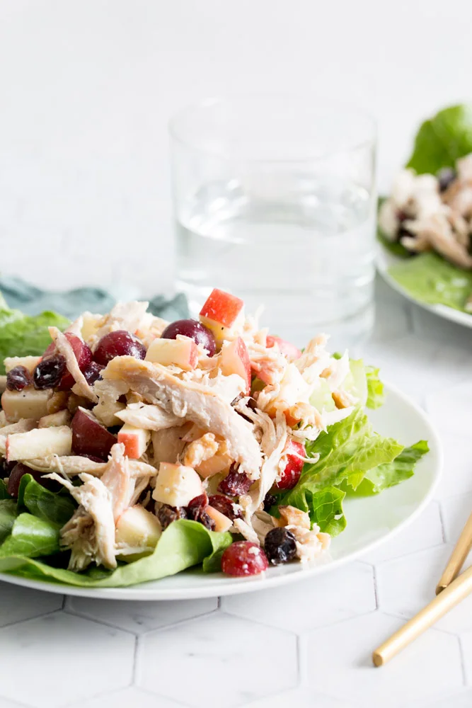 A tabletop view of chicken waldorf salad on a white plate with lettuce. A blue cloth napkin, glass of water, and gold untensils