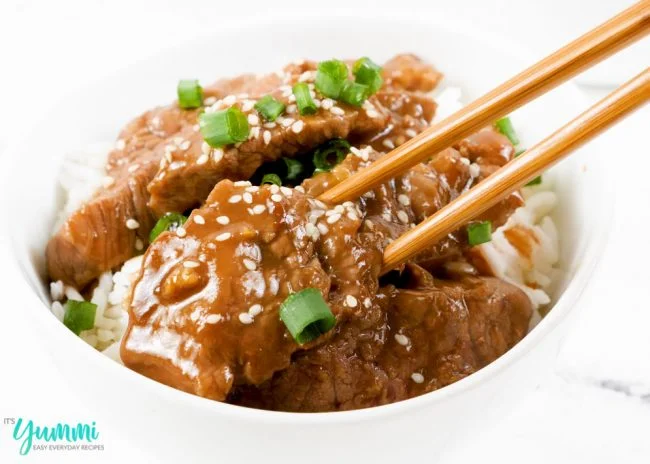 Close-up shot of chopsticks holding a piece of pf changs mongolian beef strip with scallions and sesame seeds on top