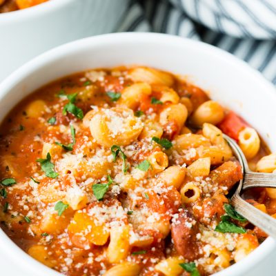 Image of: a close up of a white bowl with vegetarian minestrone soup topped with cheese and parsley. A second bowl and striped cloth are in the background.