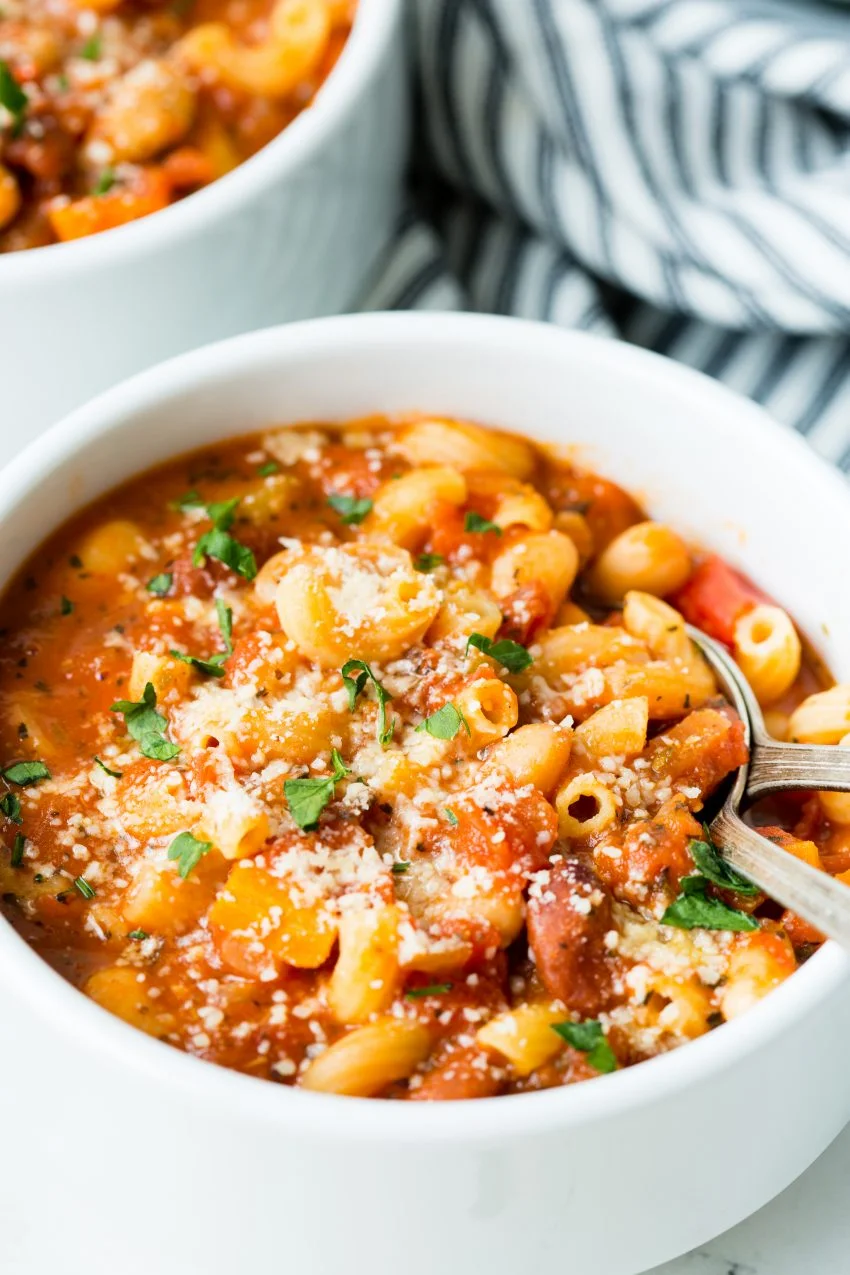 Image of: a close up of a white bowl with vegetarian minestrone soup topped with cheese and parsley. A second bowl and striped cloth are in the background.
