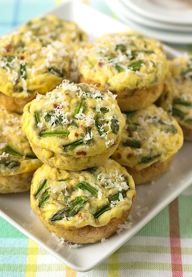 These crustless quiche muffins with protein packed quinoa are delicious for any holiday brunch, or even breakfast or lunch on the go. They're gluten-free, vegetarian, they freeze well, and they're a Weight Watcher's friendly breakfast, coming in at just 2 Smart Points per muffin! #weightwatchers #brunch #easter #glutenfree #vegetarian Quinoa quiche muffins with peppered Parmesan cheese and asparagus stacked on a white platter