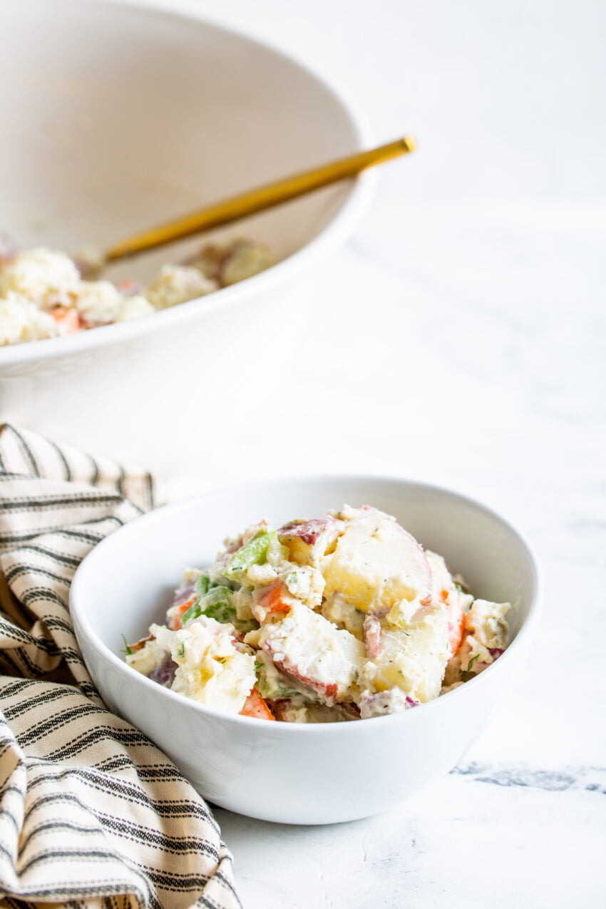 a close up view of farmers market potato salad in a white bowl, a larger serving bowl, a striped cloth napkin and gold serving utensil