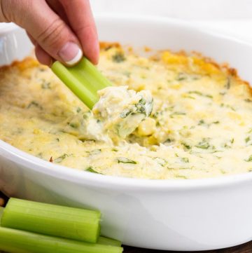 Spinach Artichoke Dip in Casserole Dish with a hand dipping a piece of celery into