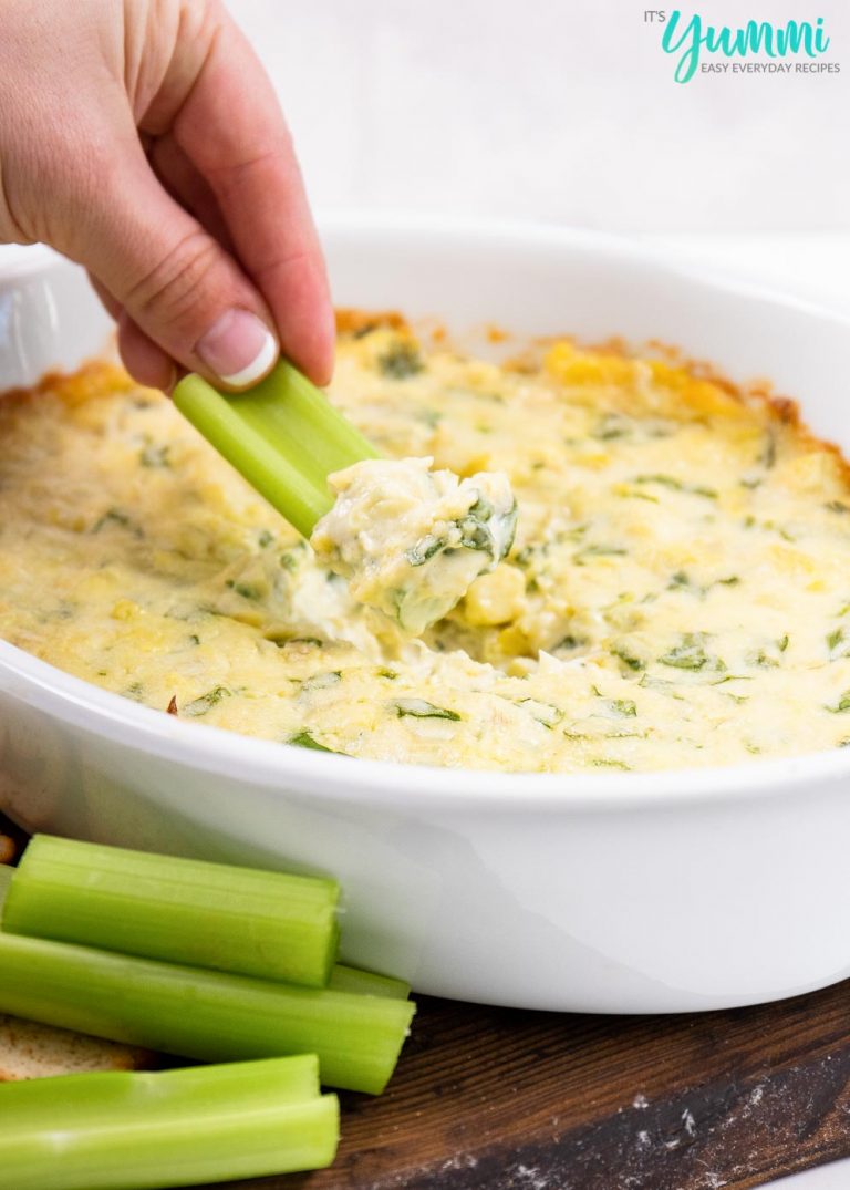 Spinach Artichoke Dip in Casserole Dish with a hand dipping a piece of celery into