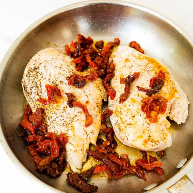 Halved chicken breasts topped with chopped sundried tomatoes and oil, in a stainless steel skillet.