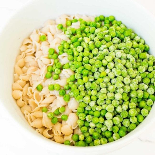 A large white bowl containing pasta shells and frozen peas.