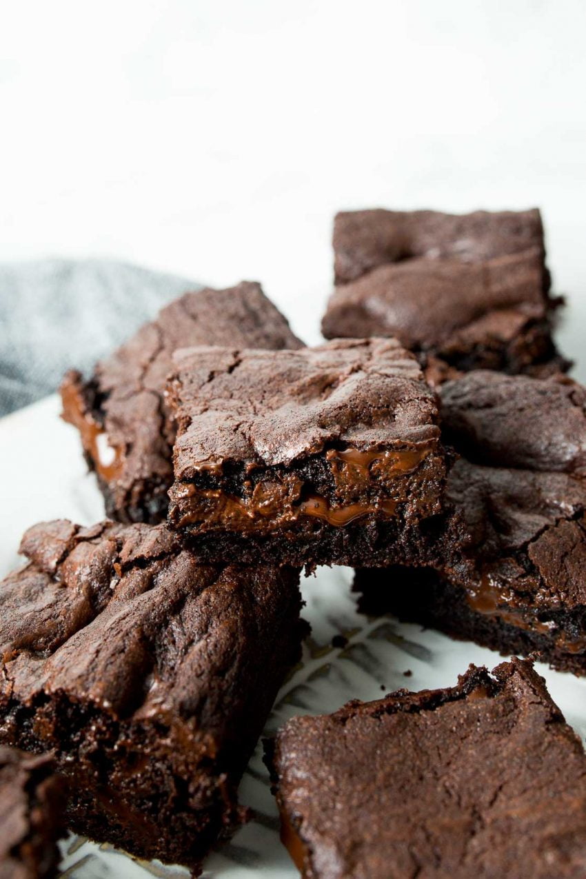 Image of: a white platter holding multiple chocolate brownies with melted fudge middles. In the background a blue table linen.