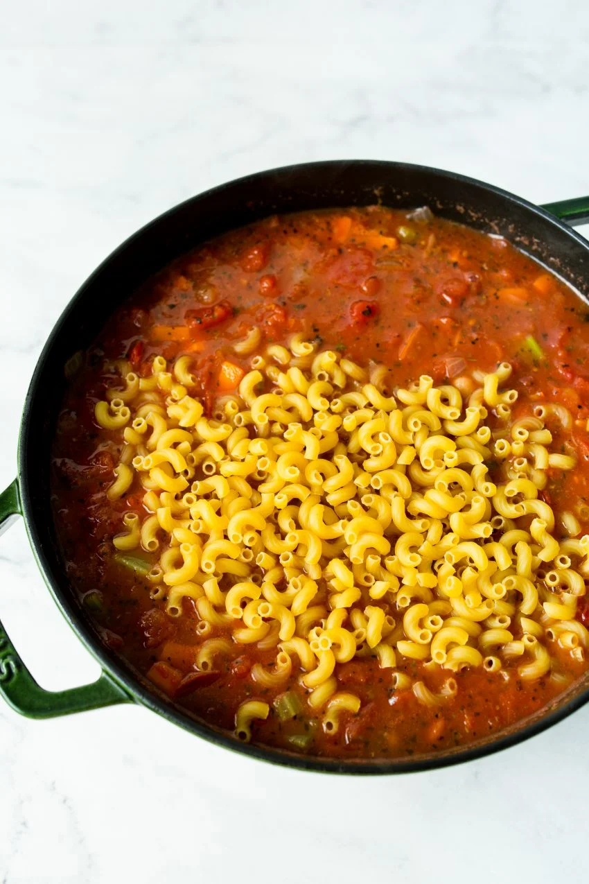 A dutch oven of minestrone soup with raw elbow macaroni.
