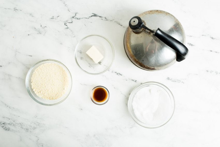 Ingredients for sweetened condensed milk on a white marble countertop: water, butter, dried milk, vanilla extract, sugar.