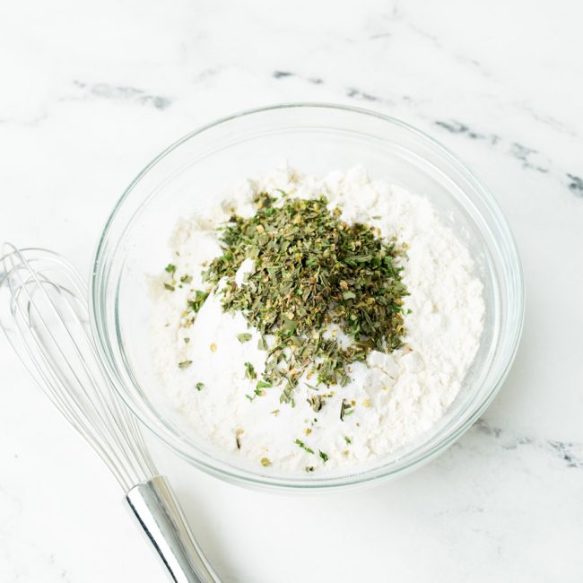 Flour, salt, baking powder, and italian seasoning in a glass bowl with a whisk to the side.