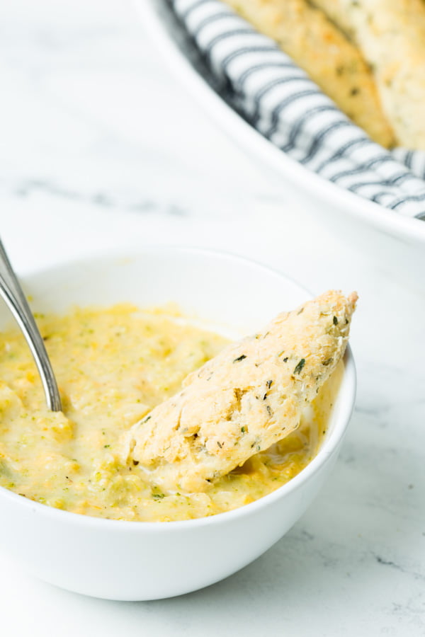 A homemade gluten-free italian breadstick dipped into a warm bowl of broccoli cheddar soup.