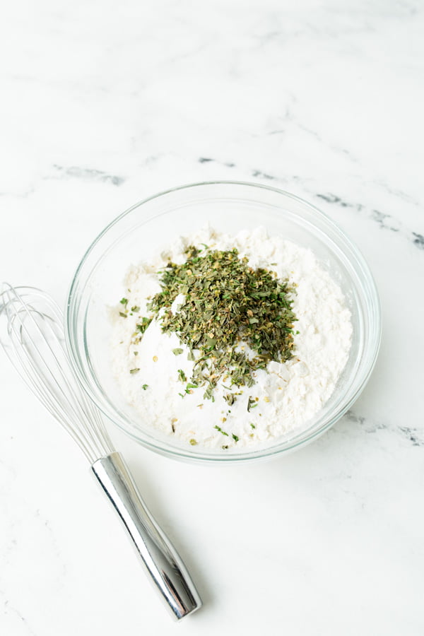 Flour, salt, baking powder, and italian seasoning in a glass bowl with a whisk to the side.