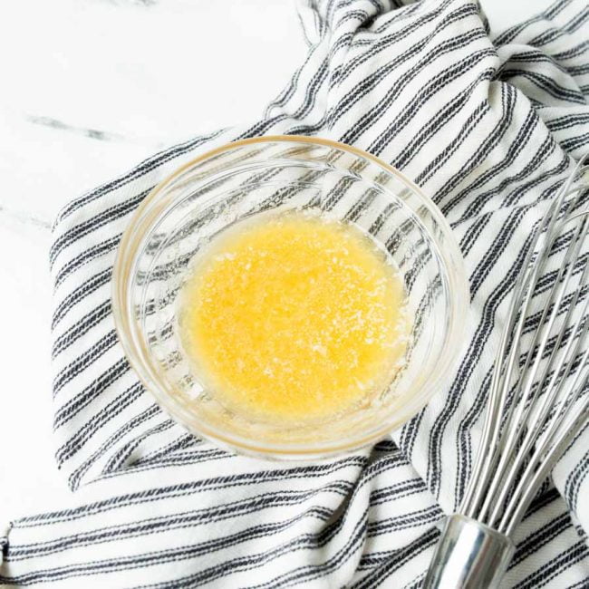 Butter and garlic powder melted in a small bowl on a white and blue cloth towel, next to a small whisk.