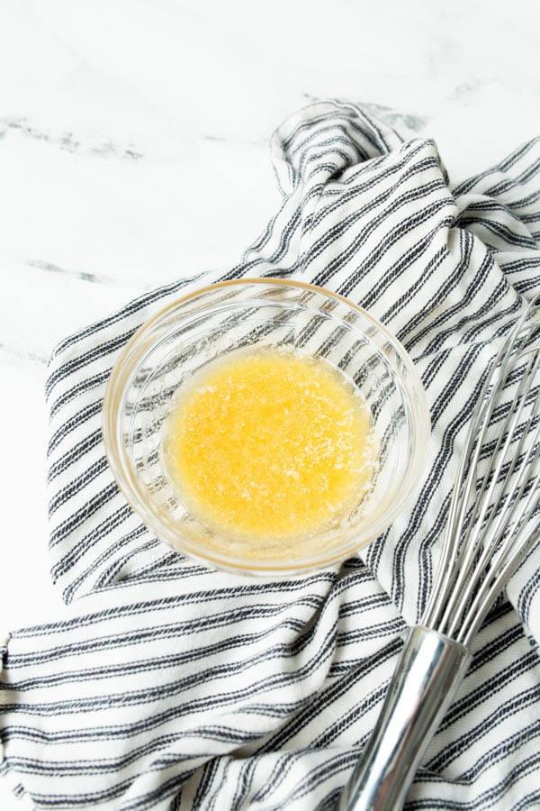 Butter and garlic powder melted in a small bowl on a white and blue cloth towel, next to a small whisk.