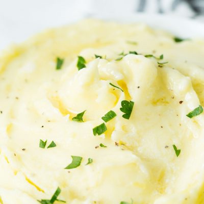 A close up image of a white bowl with perfect mashed potatoes topped with butter and chopped parsley