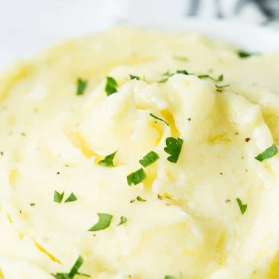 A close up image of a white bowl with perfect mashed potatoes topped with butter and chopped parsley