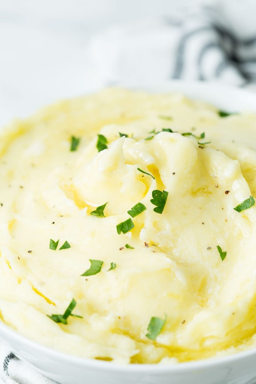 A close up image of a white bowl with perfect mashed potatoes topped with butter and chopped parsley