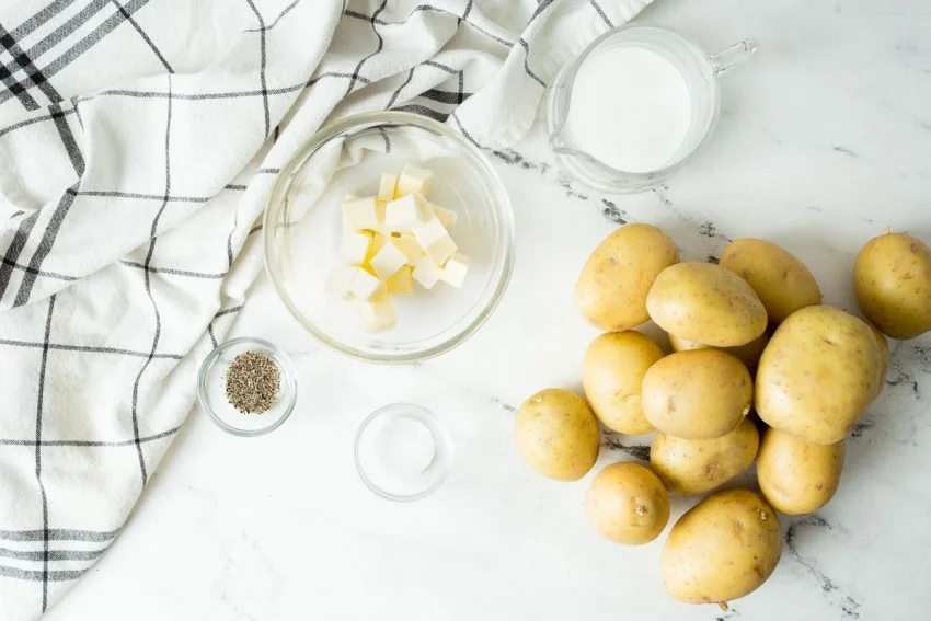 Ingredients for the perfect mashed potatoes include potatoes, milk, butter, salt, and pepper. All ingredients pictured from above on a white marble countertop.