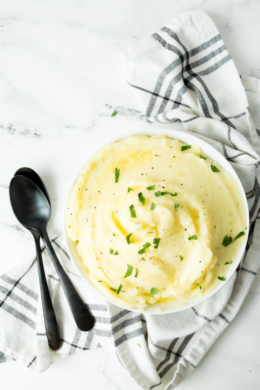 An overhead image of perfectly mashed potatoes in a white bowl next to two black spoons, topped with melted butter and chopped parsley.